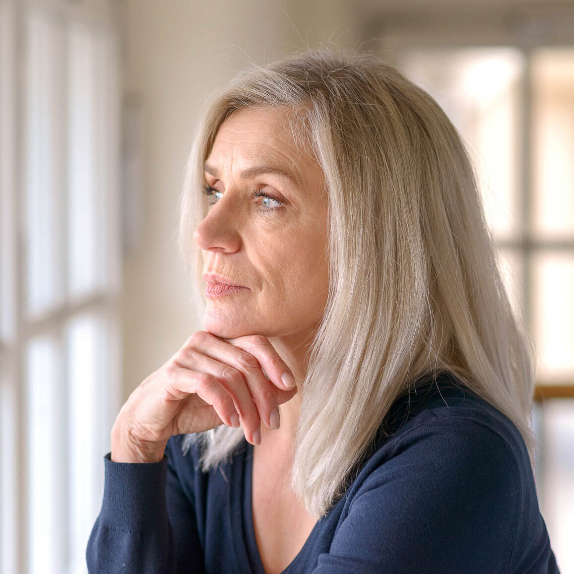 Eine Frau mit blonden Haaren greift sich nachdenklich ans Kinn und blickt aus dem Fenster